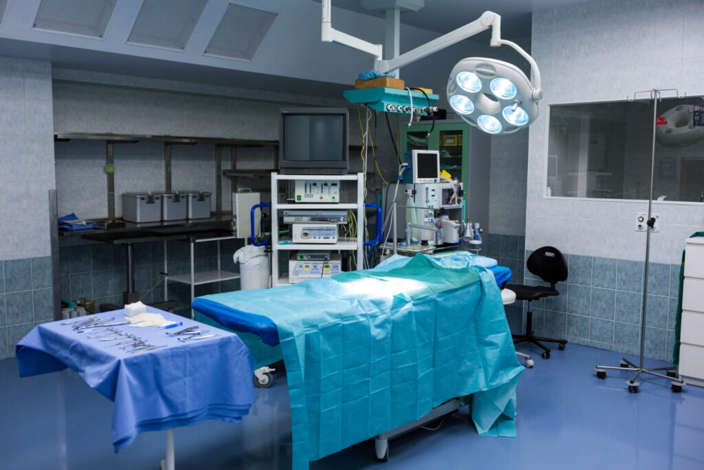 Vista de sala de cirurgia composta por uma maca, itens de bisturi e equipamentos cirúrgicos